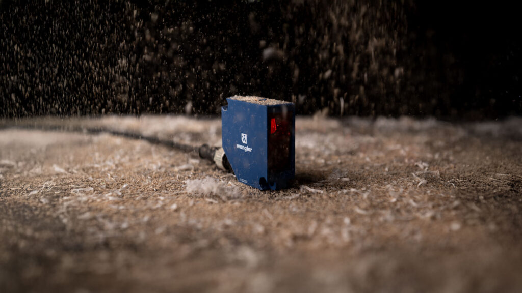 Razpršeni senzor P1PH9 podjetja wenglor stoji na površini, prekriti z žagovino, v zraku pa je prah.